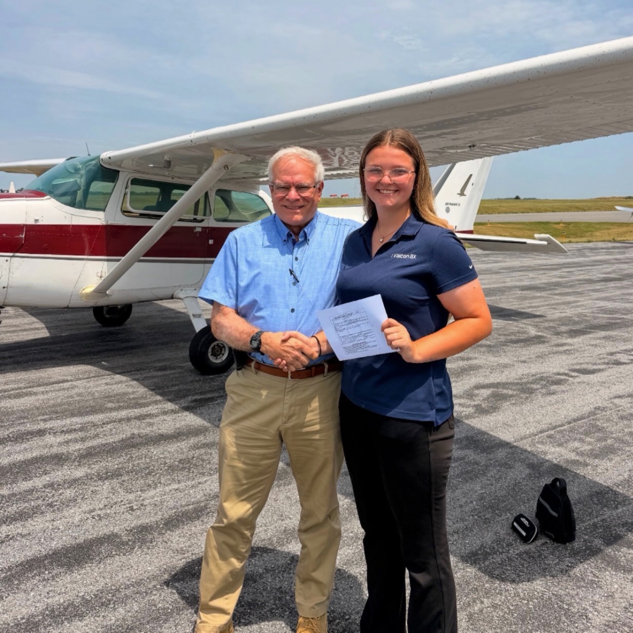 An older man and a young woman shaking hands in front of a Cessna, holding a sheet of paper