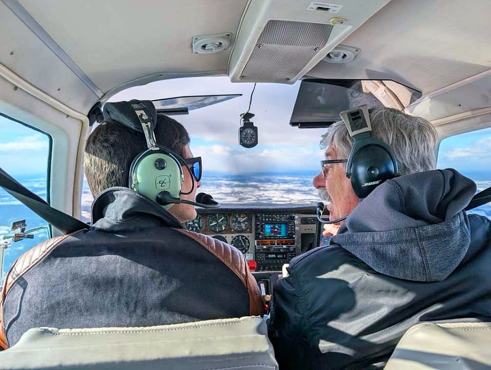 Two people in headsets seen from behind in a cockpit, looking ahead over a snowy landscape