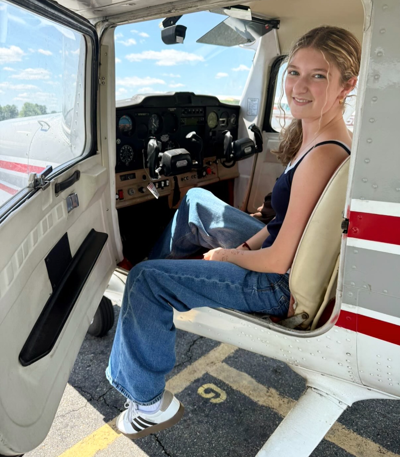 A smiling young woman sitting in the open door of a Cessna cockpit on the ramp