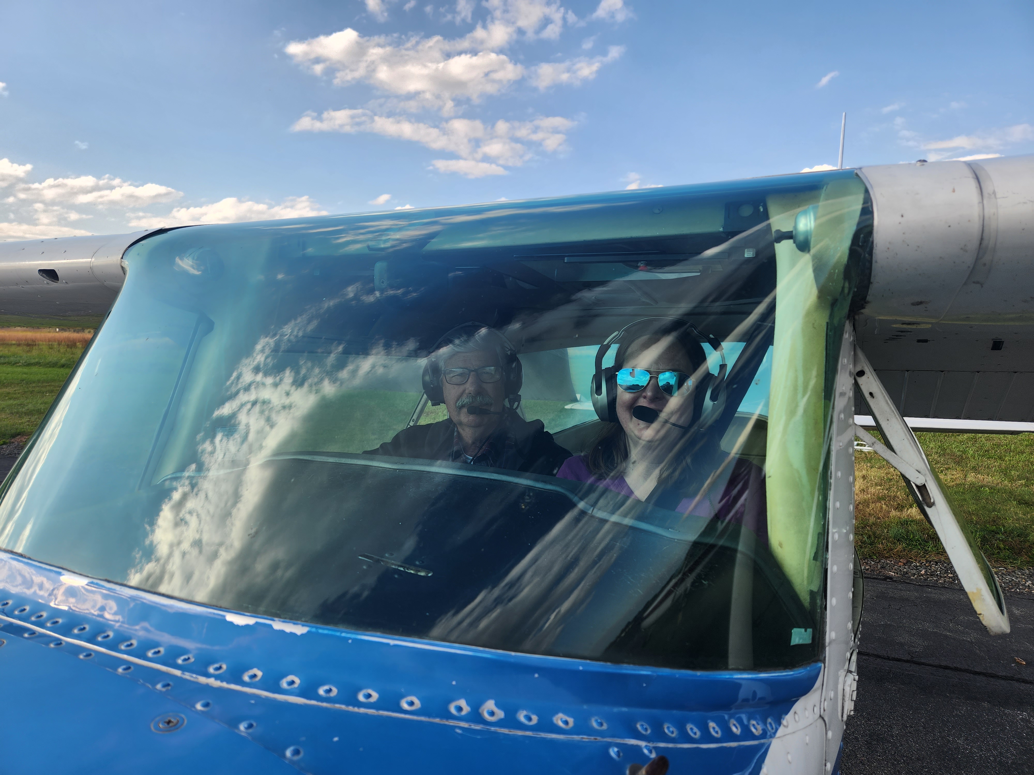 Two people wearing headsets seen through the windshield of a blue Cessna on the ground